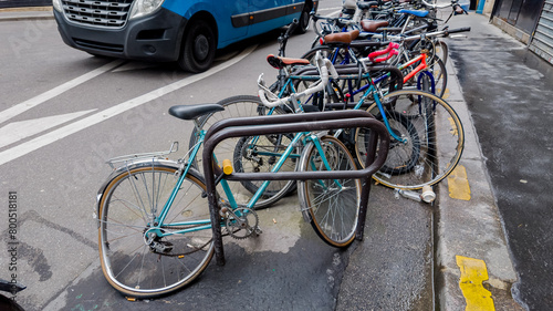 Urban bicycle parking with various bikes secured to metal racks on a city street, with a blue van passing by, depicting eco-friendly transportation