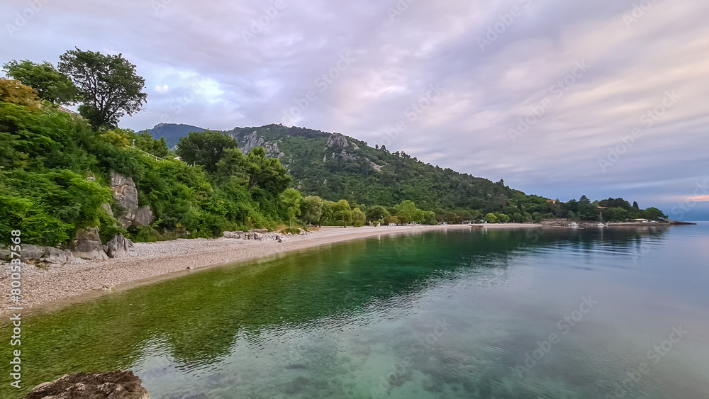 A stony beach along the shore of Medveja in Croatia. The Mediterranean Sea is calm and clear. There is a lush forest with a small town at the shore. A bit of overcast. Summer remedy. Holidays vibes.