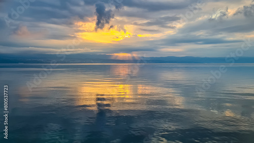 The sun setting behind mountains in Croatia. The Mediterranean Sea is calm and clear  reflecting the sunbeams. The sky is orange and covered with clouds. Many cities along the coastal line. Calmness