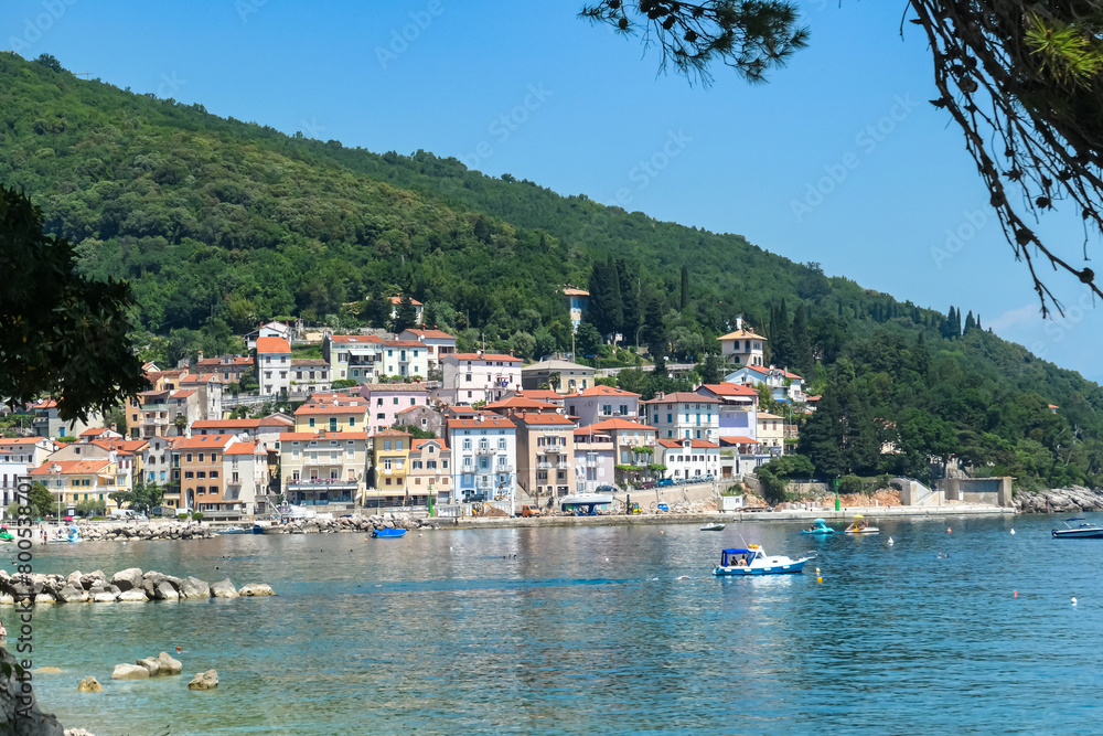 A close up on the shore along Moscenicka Draga, Croatia. There is a small town located on the shore of the Mediterranean Sea. Few boats crossing the calm sea. Green hills in the back. Summer vibes.