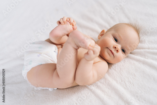 Portrait of sweet beautiful newborn baby in diapers lying on bedsheets, plays touches toes smiling looking at camera. Carefree healthy babyhood, healthcare and pediatrics care concept.