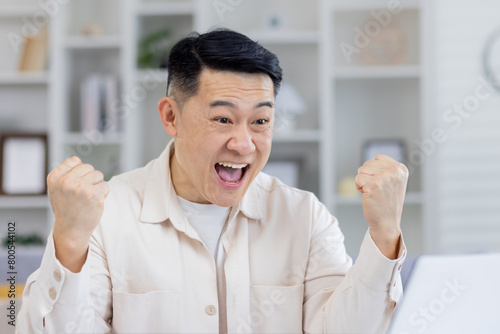 Middle-aged Asian man in a cream shirt joyously celebrating while looking at his laptop screen, evidently thrilled by good news or success at home. photo