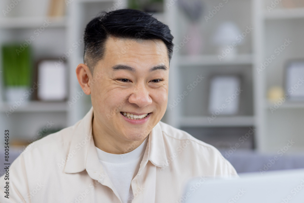 A middle-aged Asian man showing a joyful expression while interacting on his laptop in a stylishly decorated living room. Perfect imagery for themes on remote work, happiness, and technology use at A middle-aged Asian man showing a joyful expression while interacting on his laptop in a stylishly decorated living room. Perfect imagery for themes on remote work, happiness, and technology use at