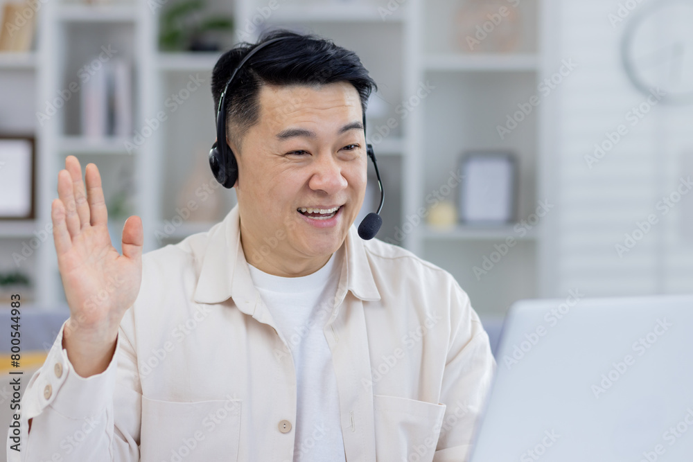 Cheerful Asian man using a headset, engaging in an online conversation from his home office. His expression is friendly and welcoming, suggesting a professional yet casual remote work environment. Cheerful Asian man using a headset, engaging in an online conversation from his home office. His expression is friendly and welcoming, suggesting a professional yet casual remote work environment.
