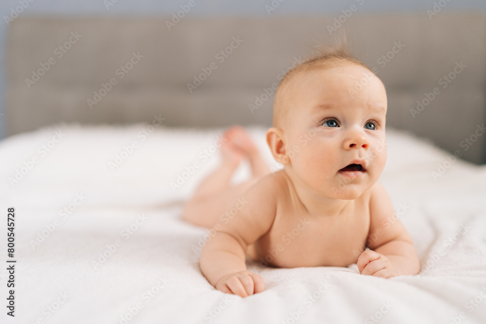 Closeup of cute little baby girl lying on stomach on bed, learning to hold head. Portrait of nice newborn naked baby looking interestingly away with open mouth. Carefree healthy babyhood concept. Closeup of cute little baby girl lying on stomach on bed, learning to hold head. Portrait of nice newborn naked baby looking interestingly away with open mouth. Carefree healthy babyhood concept.