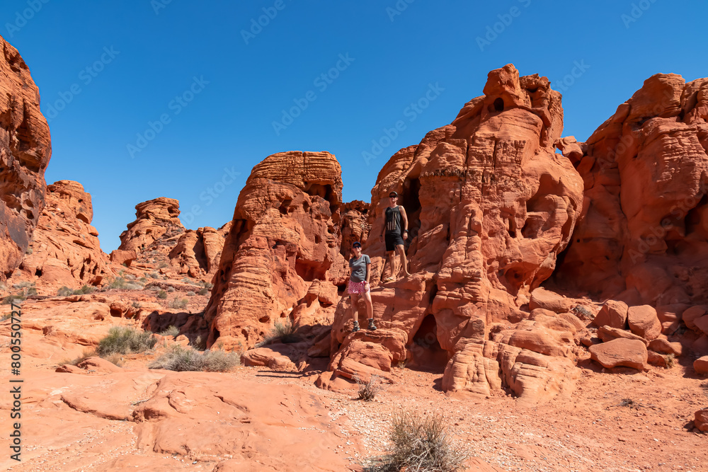 Fototapeta premium Couple at the entrance of windstone arch (fire cave) in Valley of Fire State Park, Mojave desert, Nevada, USA. Scenic view of beehive shaped red sandstone rock formations. Barren deserted landscape