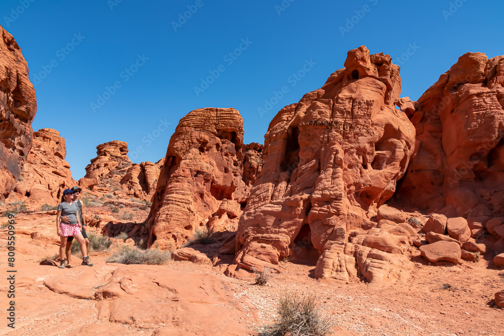 Fototapeta premium Couple at the entrance of windstone arch (fire cave) in Valley of Fire State Park, Mojave desert, Nevada, USA. Scenic view of beehive shaped red sandstone rock formations. Barren deserted landscape