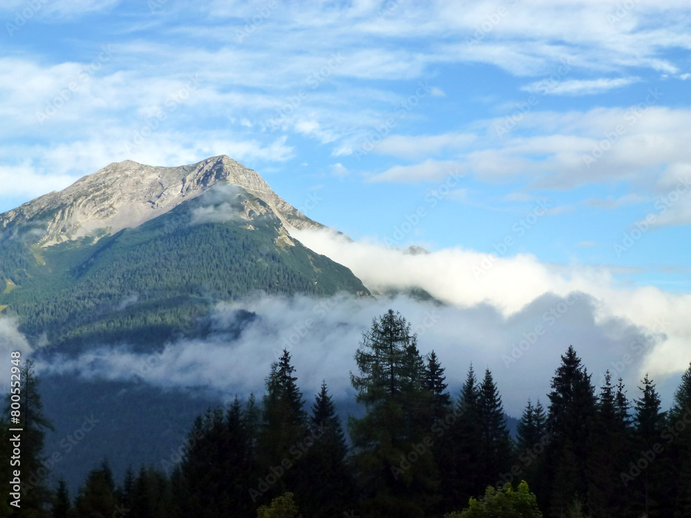 Obraz premium A picturesque view of a mountain rock overgrown with a green forest. Above them is a blue sky and white clouds