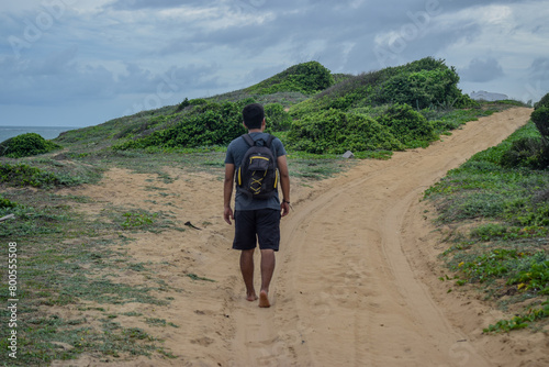 Man walking along the coast of a beach carrying a backpack on his back, between sand dunes next to the ocean, rear view
