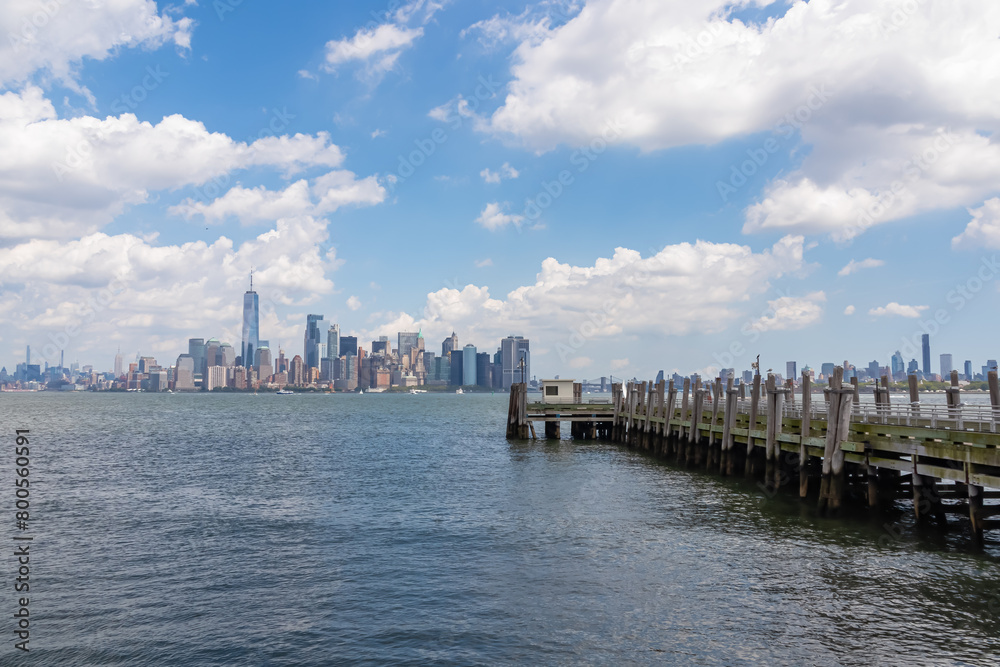 A wooden pier on the Hudson River with captivating New York urban skyline with striking and modern skyscrapers in the back, seen from The Battery Park. Think clouds above the city. Modern city.
