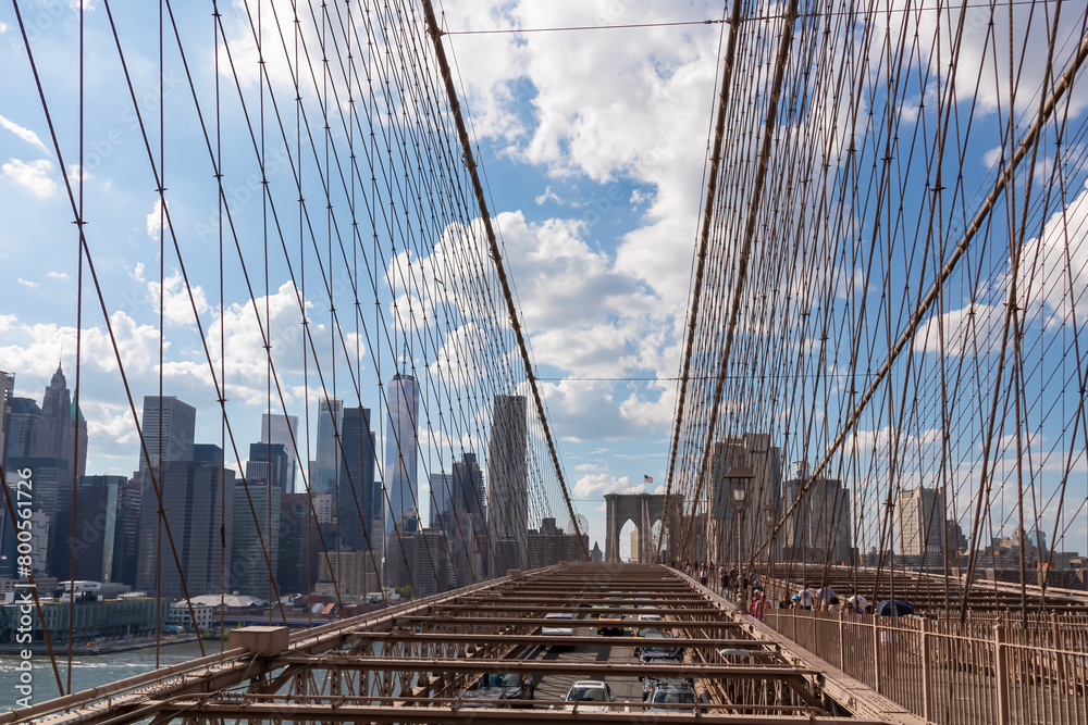 Obraz premium The gate of Brooklyn Bridge with a waving American flag on top of it contrasted with a blue sky with puffy, white clouds. Suspension bridge in New York City. Numerous skyscrapers in the back.