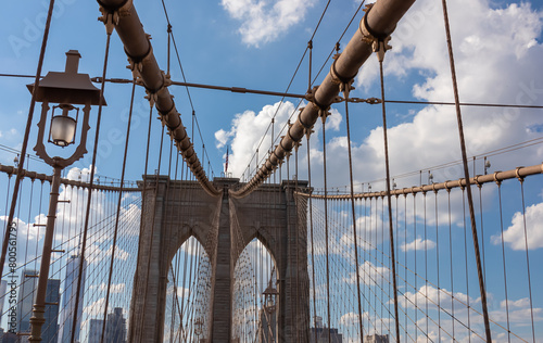 The gate of Brooklyn Bridge with a waving American flag on top of it contrasted with a blue sky with puffy, white clouds. Suspension bridge in New York City. Numerous skyscrapers in the back.