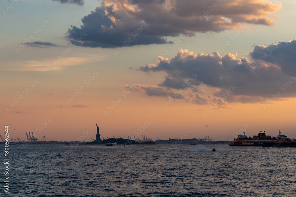 A distant view on Statue of Liberty from Battery Park. There are a lot of factories over the horizon line. Few puffy clouds on the sky. Gentle colors of sunset. Sightseeing in a big city.