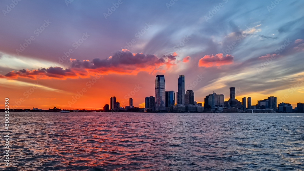 Manhattan cityscape at sunset with iconic skyscrapers reflecting on the Hudson River. Great density of skyscrapers. The sky is bursting with orange and red. Vibrant and modern city. New York at dusk.