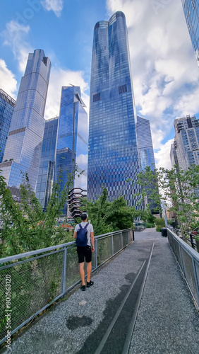 A man with a backpack enjoying the views along the famous High Lane in New York. Plenty of skyscrapers around, reflecting the blue sky and white clouds. The part is overgrown with trees on both sides