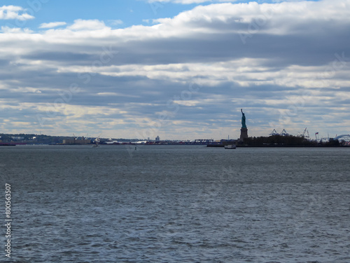 A distant view on Statue of Liberty from Battery Park. There are a lot of factories over the horizon line. Few puffy clouds on the sky. Gentle colors of sunset. Sightseeing in a big city.