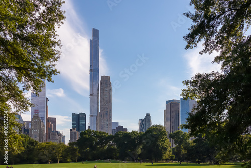 Modern cityscape of New York framed with trees with striking skyscrapers and greenery against a vibrant blue sky, seen from Central Park. The sun is shining bright. Few clouds. Summer in the city.