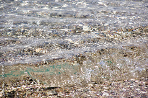 Crystal clear water rippling on rocky shoreline. Transparent water waves gently lapping over a bed of pebbles and seashells at the edge of a serene rocky beach.