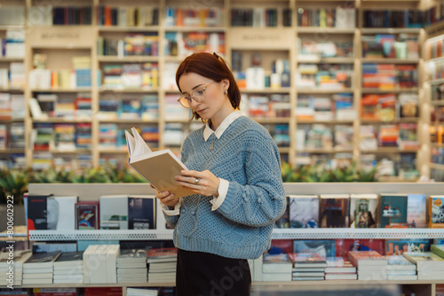 A focused young woman in glasses and a blue sweater reads a book while standing in a vibrant, well-stocked bookstore. The shelves are filled with a variety of books. photo