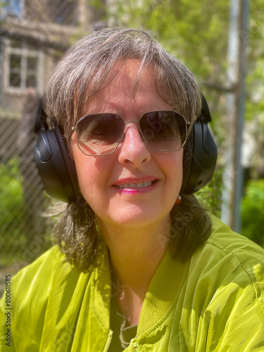 Portrait of mature, hip woman sits in a spring garden setting, wearing a green modern jacket and sunglasses, exuding confidence and style amidst the blossoming nature.