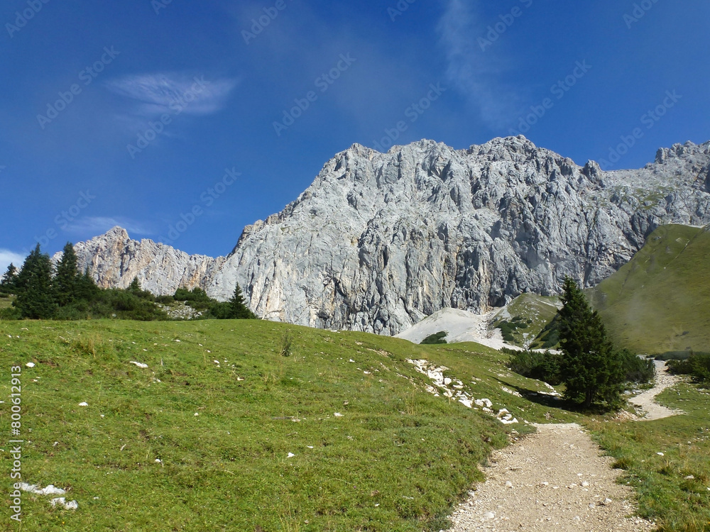 A picturesque view of a mountain slope with a dirt path and green grass. Above her is a blue sky with white clouds. View from below