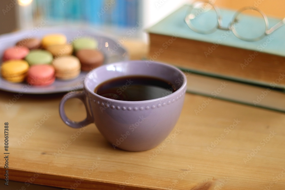 Purple plate filled with pastel macarons, cup of tea or coffee, vintage books and reading glasses on the table. Colorful bookcase in the background. Selective focus. Purple plate filled with pastel macarons, cup of tea or coffee, vintage books and reading glasses on the table. Colorful bookcase in the background. Selective focus.