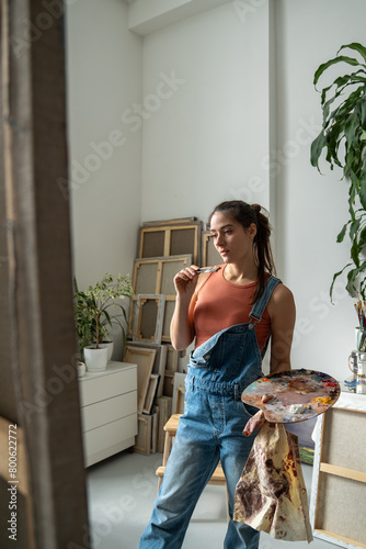 Thoughtful woman artist looks on canvas on easel pondering about creative idea for picture holds oil colour palette, paintbrush standing in art studio. Small craft business, stress relieve interests.