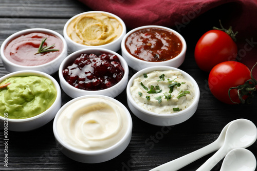 Different tasty sauces in bowls, tomatoes and spoons on black wooden table, closeup