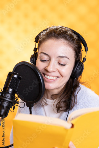 Happy narrator wearing headphones reading aloud from book into mic against yellow background. Joyous lady recording audiobook, creating engaging media content for listeners