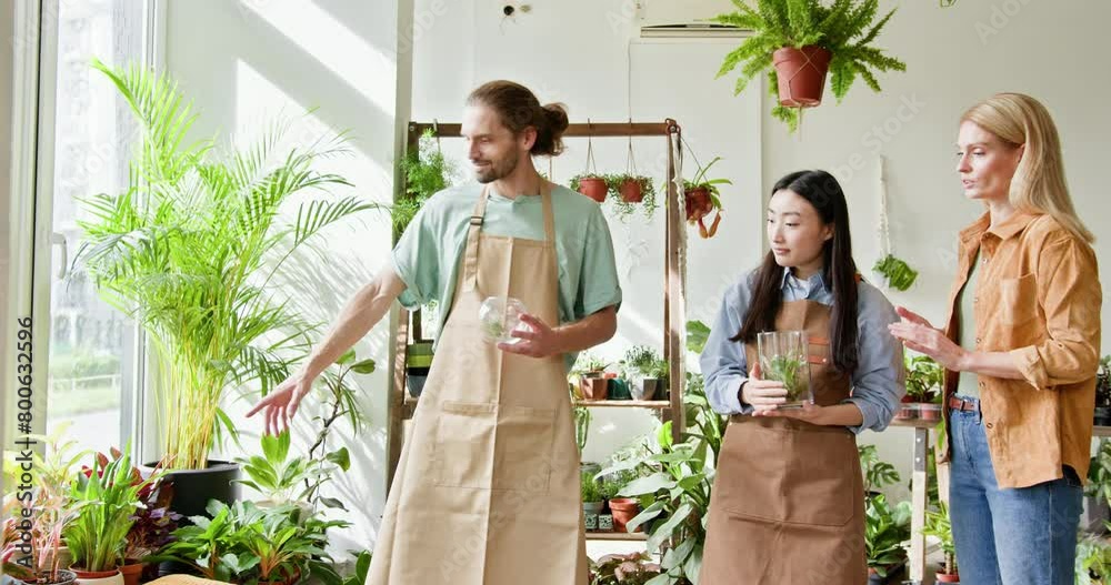 A diverse team of three, including an Asian female, collaborates in a sunlit greenhouse, sharing knowledge. People in flower shop, blond mature female client choosing a plants for her home. A diverse team of three, including an Asian female, collaborates in a sunlit greenhouse, sharing knowledge. People in flower shop, blond mature female client choosing a plants for her home.