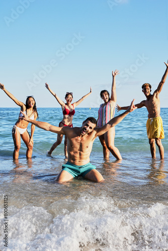 Vertical Portrait of a group of happy young people in swimming costumes posing for a photo on the shore of the beach. Diverse friends smiling and making playful gestures raising arms looking at camera © CarlosBarquero