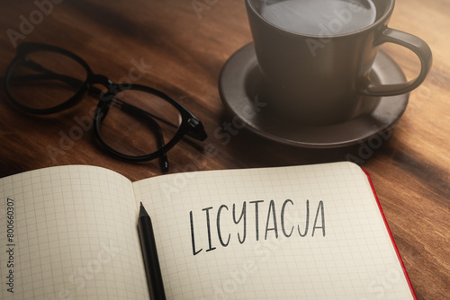  A handwritten inscription "Licytacja" on a grille of an open notebook on a wooden countertop, next to a black pencil, a cup with coffee and glasses, a flash of light. (selective focus)