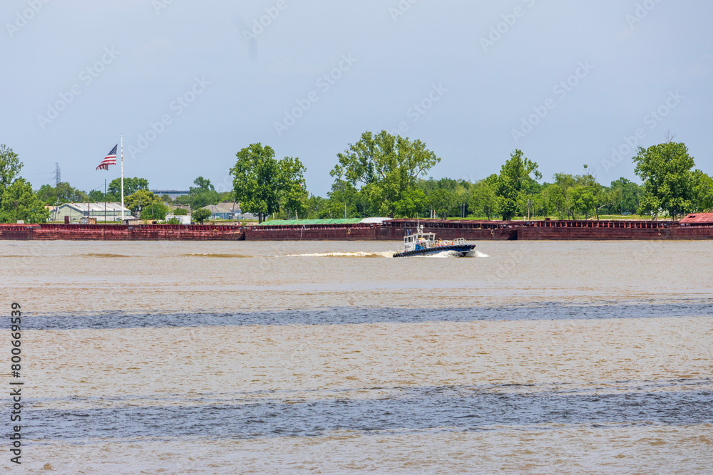 Obraz premium A motorboat sailing along the Mississippi River with an American flag flying and lush green trees, plants and grass in New Orleans Louisiana USA