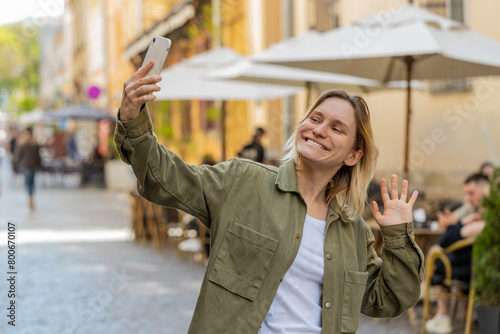 Young woman blogger taking selfie on smartphone, communicating video call online with subscribers, recording stories for social media vlog outdoors. Tourist traveler girl walking on urban city street.