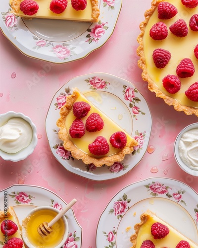 Beautiful lemon tart slices on vintage plates with fresh raspberries and honey  arranged in an elegant pattern against the soft pink backdrop of a kitchen table. 