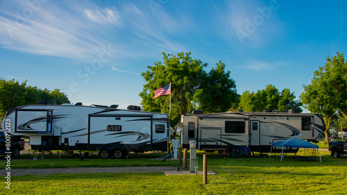 Fifth wheel trailers parked back to back at campsite on grass with trees and blue cloudy skies