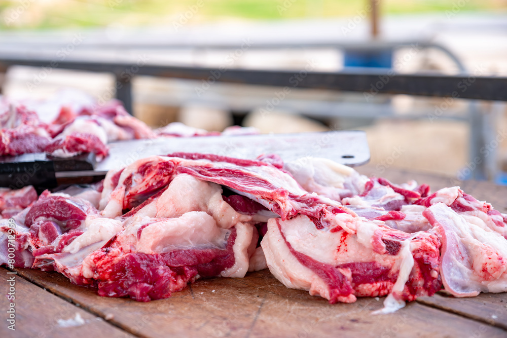 In the springtime, a bearded butcher is cutting meat at a wooden table outdoors, with sheep in the background