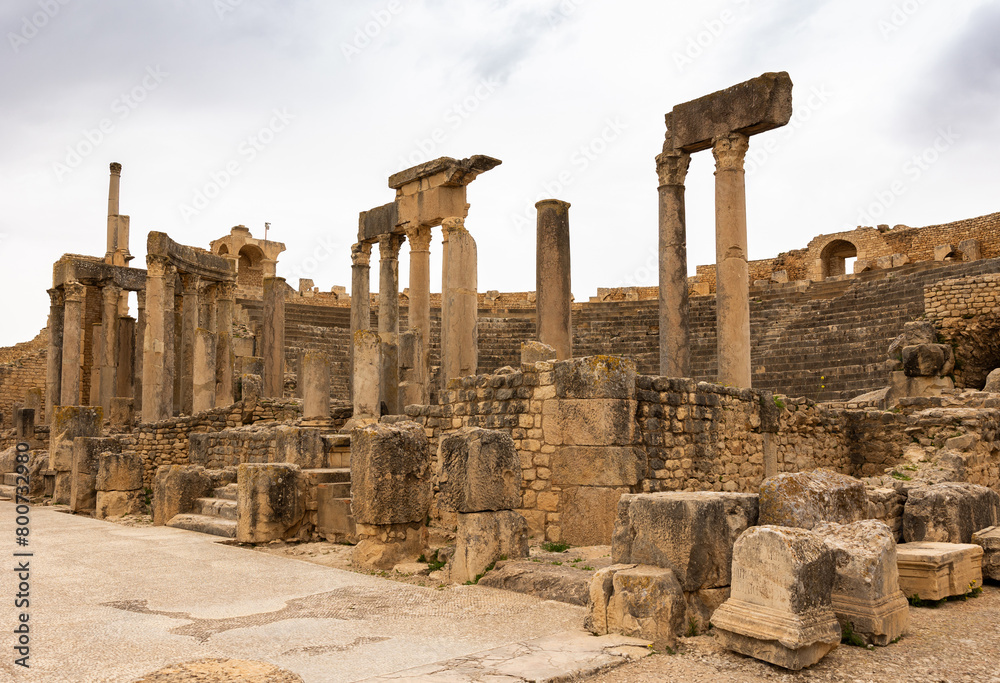 View of remains of ancient stone stage columns and seating steps of Roman theatre in Dougga against cloudy spring sky. Historical archaeological sites of Tunisia..