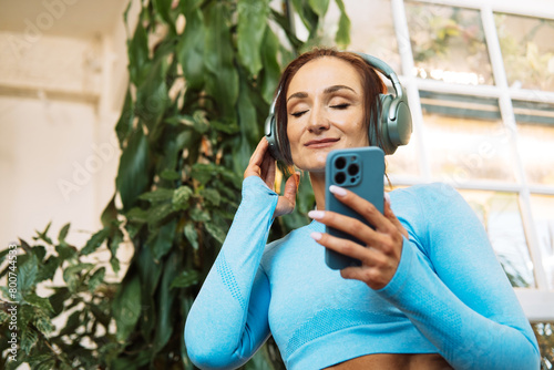 Woman wearing headphones listens to music, relaxes during Pilates, yoga training in a room with tropical plants.