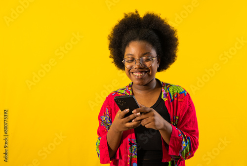 mujer joven afro sonriendo y usando lentes mientras mira su telefono movil que sostiene en sus manos  photo