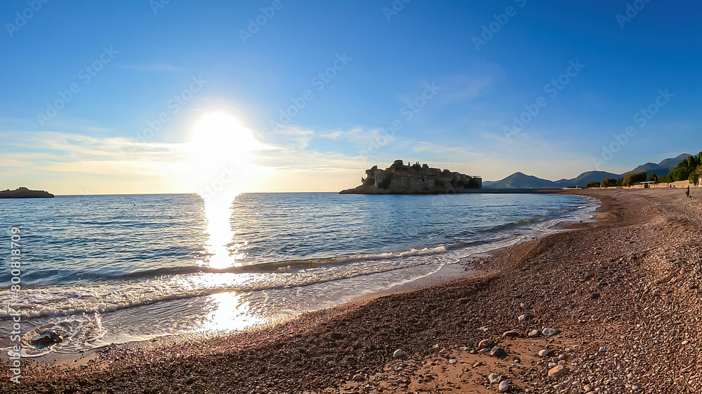 Fototapeta premium Incoming wave on sand beach with scenic view on idyllic island Sveti Stefan, Budva Riviera, Adriatic Mediterranean Sea, Montenegro, Europe. Summer vacation in luxury seaside hotel. Dinaric Alps
