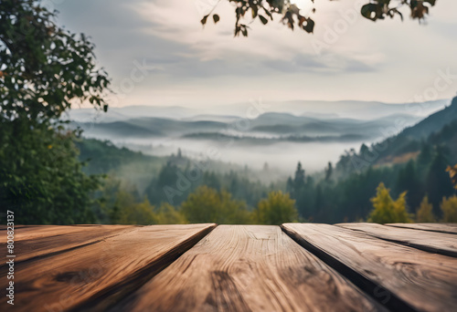 Wooden table overlooking a misty mountain landscape at sunrise, with layers of hills and a forest in the foreground.