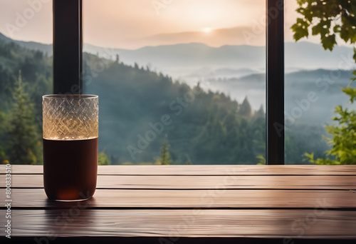 A glass of drink on a wooden table with a scenic view of a misty mountain landscape at sunrise through a window.