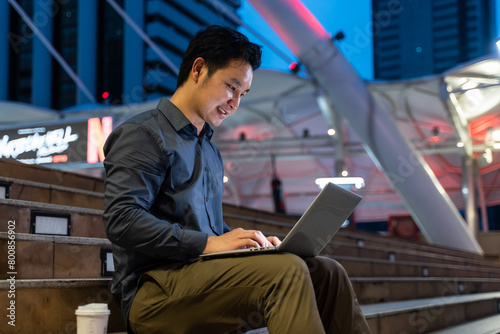 Asian young businessman sitting and working outdoors in city at night. 