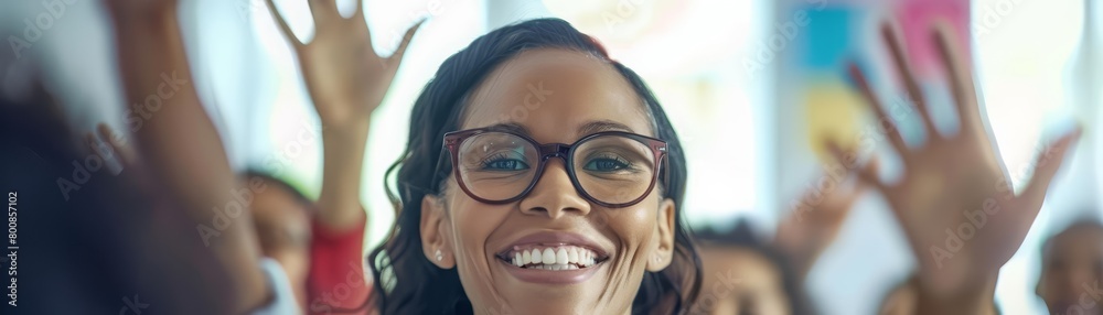 A young African-American female teacher is smiling at her students while they are raising their hands in a classroom. A young African-American female teacher is smiling at her students while they are raising their hands in a classroom.