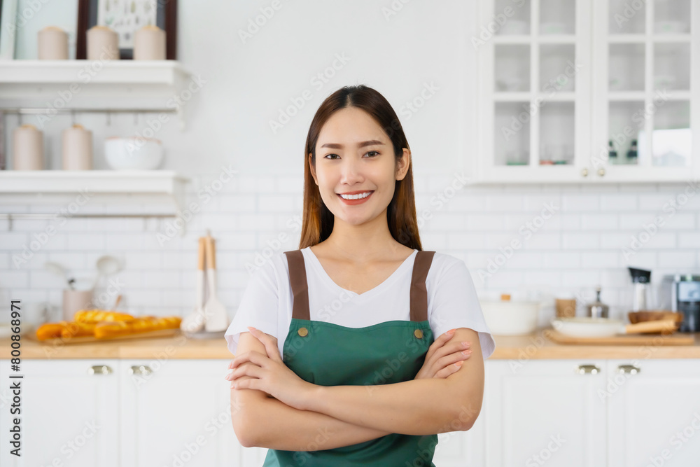 Happy young asian woman wearing apron and standing in kitchen room. Beautiful female smiling and looking at camera feeling confident Happy young asian woman wearing apron and standing in kitchen room. Beautiful female smiling and looking at camera feeling confident