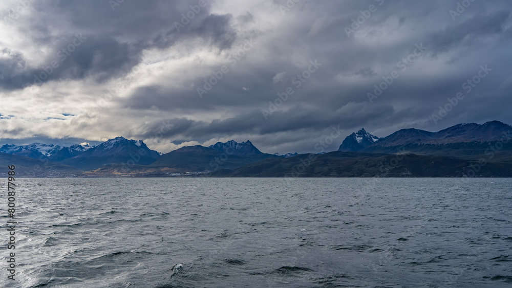 The picturesque snow-capped Andean Martial mountain range is visible from the side of the Beagle Canal. The city of Ushuaia is at the foot of the mountains on the ocean. Seabirds fly over the water