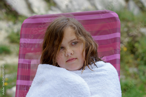 pretty eight-year-old girl sitting on a chair wrapped in a white towel after a swim at the beach, looking cold.