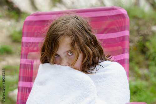 pretty eight-year-old girl sitting on a chair wrapped in a white towel after getting out of the water, looking cold.