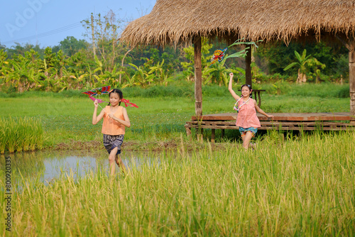 Happy Asian Thai group of boys and girls wear traditional colorful farmer outfits and run with flying kites on a green field.